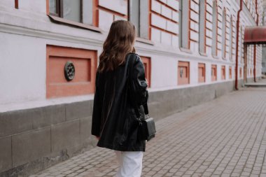 Siyah deri ceketli, beyaz pantolonlu, beyaz duvarın önünde yürüyen tanınmayan bir kadın. Şehir tarzı ve sokak modası. Kıvırcık saçlı, günlük kıyafetli bir kız.