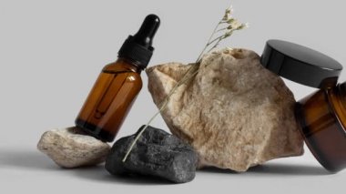 Damlatan cam pipet şişesinin hareketli resmi, bir kavanoz krema, çiçek dalları, kayaların üzerinde denge, gri arka plan. Serum, podyumda asit. Organik, doğal kozmetik. Güzellik, cilt bakımı
