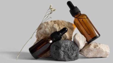 Taşların üzerinde duran damlatan cam pipet şişelerinin hareketli resmi, çiçek dalı, gri arka plan. Serum, podyumda asit. Sıfır atık cam. Organik, doğal kozmetik. Spa, güzellik, cilt bakımı