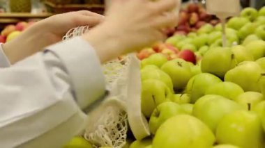 Kadın eli, elmaları topluyor, seçiyor, beyaz ağ torbasında katlıyor. Süpermarkette, markette çevre dostu alışveriş. Yeniden kullanılabilir pamuk iplik torbasında meyveler. Sıfır atık ve plastiksiz kavram.