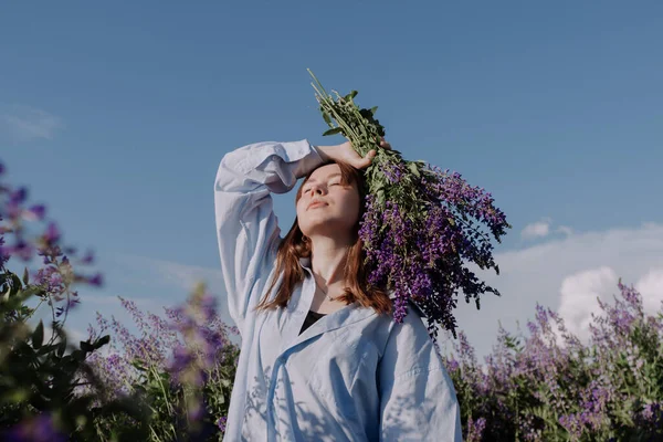 Smilende ung kvinde i skjorte holder buket lilla blomster stående alene blandt feltet foran blå himmel nyder af sollys på gyldne time. Sommertøj. Pige med blomster og lukkede øjne - Stock-foto