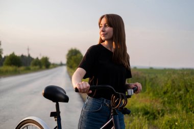 Kırsalda bisikletle tek başına duran genç bir kadın sola bakıyor. Yaz aktivitesi, sağlıklı yaşam tarzı, egzersiz, spor, fitness. Doğadaki insan. Bisiklete binmek. Dışarıda eğleniyoruz..