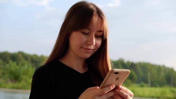 Smiling young woman in black t-shirt using smartphone, scrolling feed at countryside. Using device. Girl looking down and texting on smartphone. Chatting, messaging, reading. — Stock video