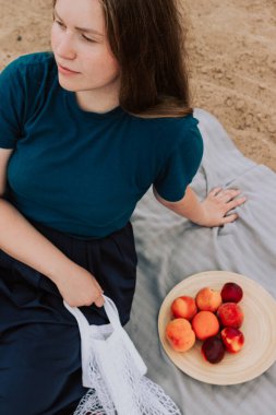 Kumda oturan meyveli beyaz ağ torbası tutan bir kadın. Çevre dostu piknik, yeniden kullanılabilir alışveriş çantası. Şeftali, pamuk iplik torbasında portakallar. Sıfır atık ve plastiksiz kavram.
