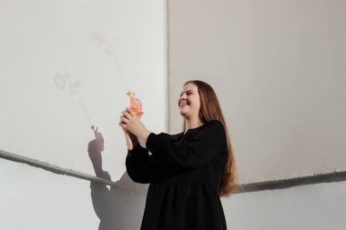 Beyaz duvar arkasında sabun köpüğü oyuncağı kullanarak siyah elbiseli gülümseyen genç bir kadın. Açık havada yaz etkinliği. Şehirde eğleniyoruz. Mutluluk, Pozitif Duygu. Günlük kıyafet.