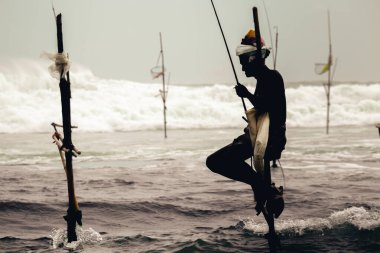 Weligama, Sri Lanka - 07: 26: 2020: Yaşlı balıkçı oturmuş, akşam siluetinde balık tutarken, okyanus gelgitleri arka planda, sabır ve bekleme oyununda kaza yaparken tahta bir direğe bindi.