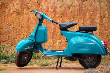 Eski model motosiklet kil duvara karşı sergileniyor. Açık mavi renk ve iki koltuklu, depo için kompartımanlar, ön tarafta yuvarlak far..