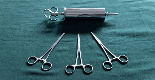 Stainless steel syringe and straight Hemostatic Forceps scissors on a green-clothed table top close up. The ear syringe is used for ear wax debris removal