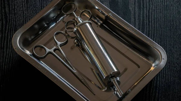 Ear syringe for ear wax debris removal and straight Hemostatic Forceps in a stainless steel container on a wooden table overhead view close-up photograph.