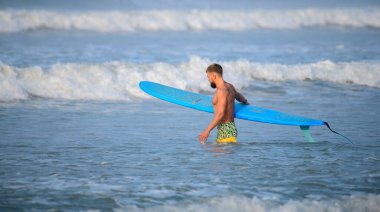 Weligama, Sri Lanka - 04 14 2022: Sığ sularda mavi bir uzun tahta taşıyan yakışıklı adam.