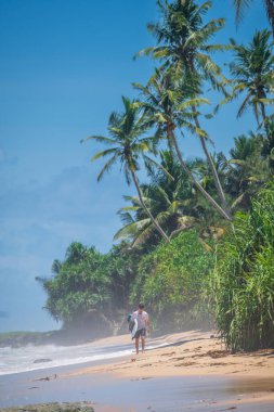 Weligama, Sri Lanka - 05 04 2023: Tahtasını taşıyan sörfçü tropikal dalgaları aştıktan sonra altın kıyılarda yürüyor. Palmiyeler ve bereketli sahil yeşillikleri sahili çevreleyecek.