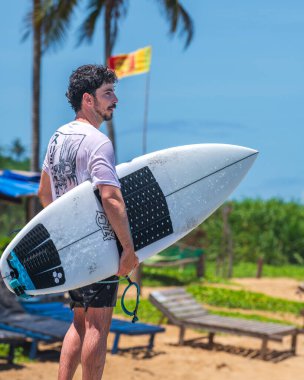 Weligama, Sri Lanka - 05 04 2023: Arka planda dalgalanan Sri Lanka bayrağına bakan beyaz ıslak tişört giyen sörfçü. Arkadan çekilmiş..