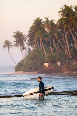 Madiha, Sri Lanka - 02: 24 2025: Bir erkek sörfçü beyaz bir kısa tahtayla güvenli bir şekilde sörfün içine girer. Sabah gökyüzü ve tropikal bir arka planda palmiye ağaçları