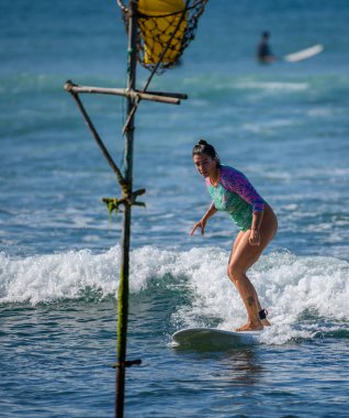 Ahangama, Sri Lanka - 02: 12: 2025: Dişi sörfçü, geleneksel bir balıkçı oltasının yanında, suyun üzerinde sorunsuzca süzülerek, odaklanmış ve kontrollü bir şekilde hafif bir dalgaya biniyor.