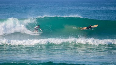 Ahangama, Sri Lanka - 02: 13 2025: Bir erkek sörfçü, hız ve kontrol için çömelmiş bir turkuaz dalganın içinde sörf yapıyor. Yakınlarda, bir kadın klasik bir sörf tahtasıyla kürek çekiyor.