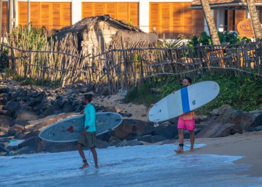 Ahangama, Sri Lanka - 02 14 2025: İki erkek sörfçü dalgalara girmeden önce sörf tahtalarını tutuyor. Arka planda engebeli kayalar ve istiflenmiş tahtalar