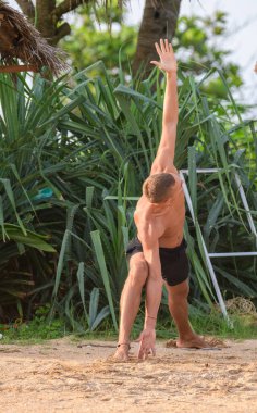 Zayıf yapılı genç bir atletik sadece siyah şort giyerek kumda derin bir esneme hareketi yapar. Vücudu bir kolu yukarı uzanmış, odaklanmayı ve esnekliği gösteriyor.