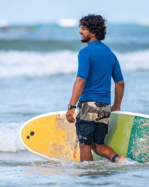 Weligama, Sri Lanka - 05: 29: 2023: Kıvırcık saçlı yakışıklı sörfçü çocuk kumlu bir sahilde dururken renkli bir sörf tahtası tutuyor.