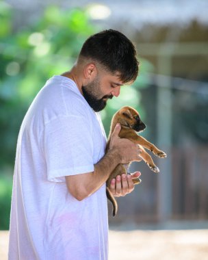 Weligama, Sri Lanka - 05 29 23: A man with a beard wearing a loose white t-shirt and orange shorts smiles warmly while holding a small brown puppy in his hands