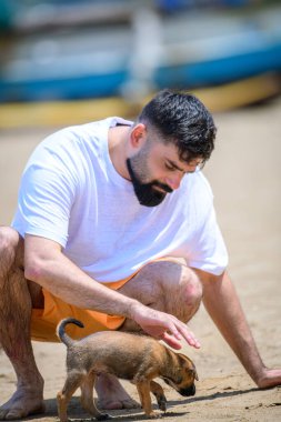 Weligama, Sri Lanka - 05 29 23: Handsome man petting a cute stray dog puppy on the sandy beach