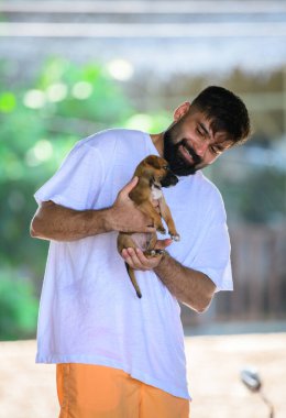 Weligama, Sri Lanka - 05 29 23: A man with a beard wearing a loose white t-shirt and orange shorts smiles warmly while holding a small brown puppy in his hands