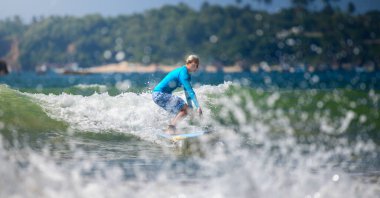 Weligama, Sri Lanka - 01 10 2025: Dişi bir sörfçü odaklanmış denge ile küçük bir dalgaya biniyor, ön planda parlak okyanus köpüğü