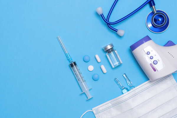 Ampoule, vial, syringe under white protective mask and non contact thermometer, stethoscope on blue background. Vaccination concept, disease prevention. Health care, Coronavirus vaccine development.