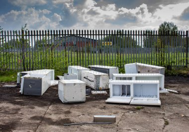 Several discarded fly-tipped fridges and freezers dumped on concrete ground in front of spiked security fence, Clayton, Manchester, UK. clipart