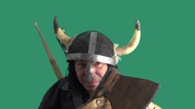 Ağır çekim. Yeşil arka plan. Bir ok Viking savaşçısının tahta kalkanına çarpıyor. Boynuzlu ve elinde balta olan kasklı olgun bir adam.