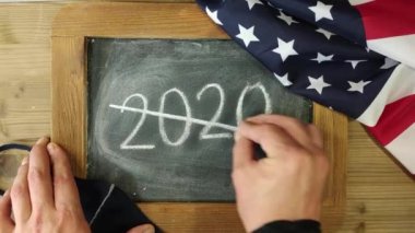 Tahtaya tebeşirle 2020 yazısını tahtanın kenarları boyunca Amerikan bayrağı ve siyah maskesini virüsten koruyan tahtalarla insan eli çiziyor.