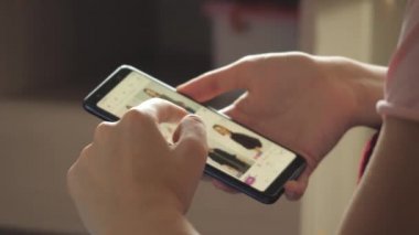 Online Mağaza Giysilerindeki Mallara Bakıyor. Moda kıyafetlerini doğrudan Smartphone 'dan al. Evdeki Kadın Oturma Odasındaki Koltukta Uzanıyor İnternet Dükkanından Akıllı Telefon Satın Alıyor