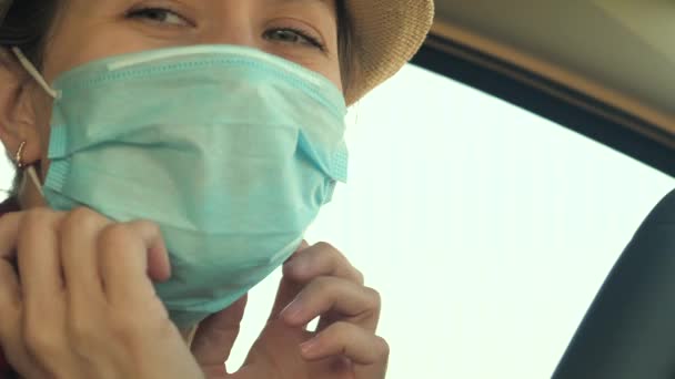 Glad pige sætter på en medicinsk maske på hendes ansigt og smiler mens du kører i bilen. Pandemi. Livet under en epidemi. Covid-19. Coronavirus beskyttelse. Rejs alene. Nærbillede – Stock-video