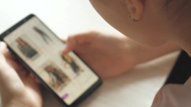 Online Mağaza Giysilerindeki Mallara Bakıyor. Moda kıyafetlerini doğrudan Smartphone 'dan al. Evdeki Kadın Oturma Odasındaki Koltukta Uzanıyor İnternet Dükkanından Akıllı Telefon Satın Alıyor — Stok video