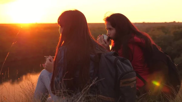 Viajantes do sexo feminino sentam-se no alto da montanha e bebem chá em uma caneca olhando para o pôr do sol. Conquistar novos lugares juntos. Fim de semana ativo. Mulheres livres turistas estão à procura de aventura na vida — Fotografia de Stock