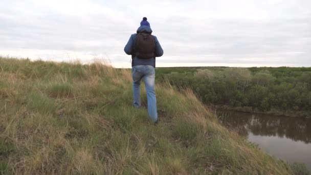 En man reser med en ryggsäck på ett berg vid floden. Han letar efter äventyr. En helg på en sportvandring ensam. Vila på naturen. Turistutforskning i området — Stockvideo