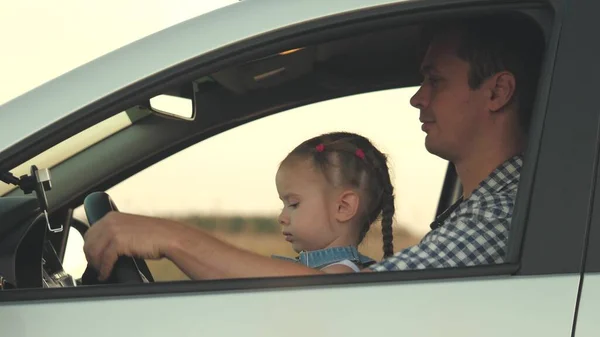 Dad teaches his little daughter to turn steering wheel while sitting in his car in drivers seat. father travels with children by car. driver and kid are driving. happy family and childhood concept