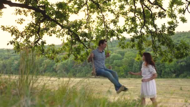 Lycklig kvinna rullar en man på en gunga och ler. Romantiska relationer mellan människor. Tillbringa tid tillsammans. Lagarbete. Sommarpromenader i naturen. Weekend livsstil — Stockvideo