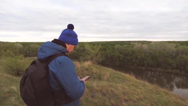 Een toerist is op zoek naar een online kaart in zijn telefoon. Reizen met een smartphone en het internet. Sociale media zijn overal. Een man reist met een rugzak op een berg bij de rivier. De man is — Stockvideo