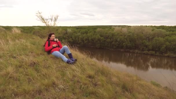 En ung flicka resenär sitter ensam på berget och dricker hett te från en mugg. Kvinna turist med en ryggsäck vilar på en weekend vandring. Vädret i naturen. Han är på semester. Ensamhet — Stockvideo