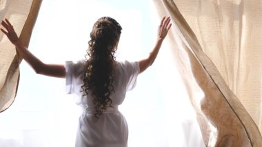 Sabahın sıcak ışığı için perdeyi çekiyorlar. Ağır çekim. Yatak odasında perdeleri açan genç bir kadın. - Yakın çekim. Sabahları eller pencerenin perdesini açar