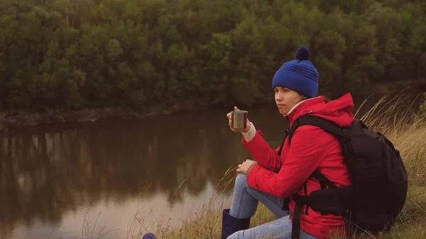 Een jonge meidenreiziger zit alleen op de berg en drinkt warme thee van een mok. Vrouw toerist met een rugzak rustend op een weekendwandeling. Winderig weer in de natuur. De man is op vakantie. Eenzaamheid — Stockfoto