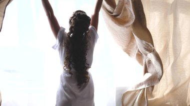 Sabahın sıcak ışığı için perdeyi çekiyorlar. Ağır çekim. Yatak odasında perdeleri açan genç bir kadın. - Yakın çekim. Sabahları eller pencerenin perdesini açar