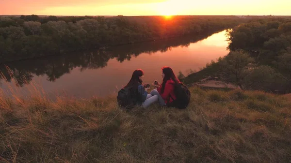 Viajantes do sexo feminino sentam-se no alto de uma montanha junto ao rio ao pôr-do-sol e bebem chá de uma caneca. Vai acampar com um amigo no fim de semana. Exploração turística da área da natureza. Livre-se de todos os dias — Fotografia de Stock