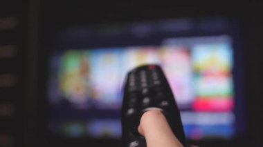 Kadın uzaktan kumandayla internet kanallarını seçiyor, yakın plan. Televizyonu modern bir uzaktan kumandayla kontrol eden kişi. Kız akıllı TV izler ve siyah uzaktan kumanda kullanır. Bulanık TV kaydırma sayfaları