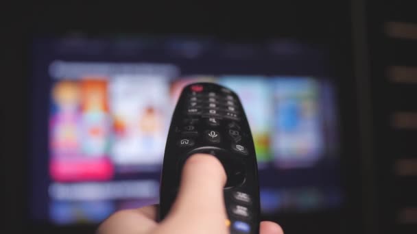 Mujer mano selecciona canales de televisión por Internet con control remoto, primer plano. La persona controla la televisión usando un moderno control remoto. Chica mira televisión inteligente y utiliza el control remoto negro. Desenfocar páginas de rollos de televisión — Vídeos de Stock