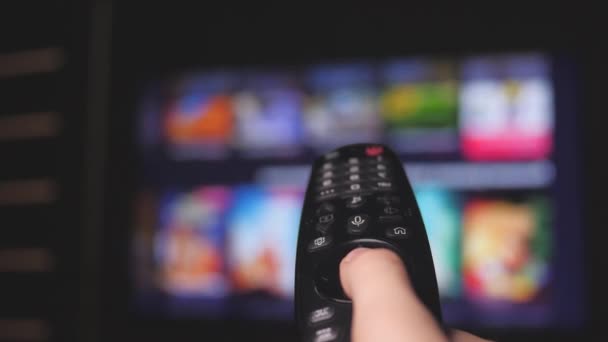 Kadın uzaktan kumandayla internet kanallarını seçiyor, yakın plan. Televizyonu modern bir uzaktan kumandayla kontrol eden kişi. Kız akıllı TV izler ve siyah uzaktan kumanda kullanır. Bulanık TV kaydırma sayfaları — Stok video