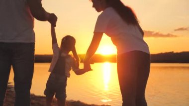 Gün batımında anne ve babanın elini tutan küçük kız. Mutlu aile hayatı konsepti. Anne ve babasıyla birlikte uçan bir çocuk. Anne ve baba çocukla yürüyüşe çıktılar. Harca