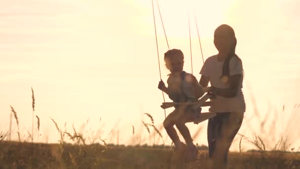 Parque de atracciones concepto de ensueño. Chica feliz balanceándose en un columpio en el parque al atardecer. niño juega con el oscilación de madera, sueño de la niñez, vuelos del piloto del avión al cielo. Una familia feliz — Vídeo de stock