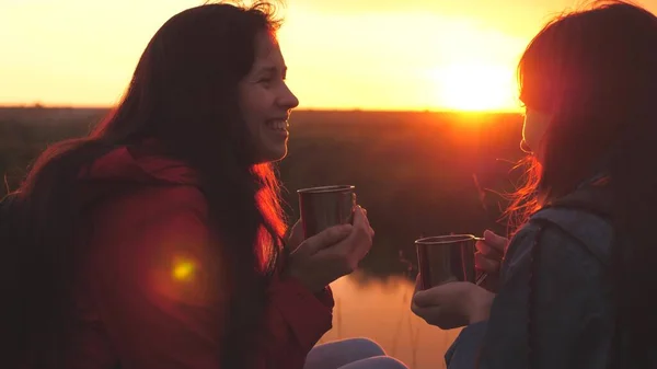 Female travelers sit high on a mountain by the river at sunset and drink tea from a mug. Go camping with a friend on the weekend. Tourist exploration of the area of nature. Free yourself from everyday