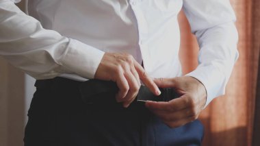 Akıllı bir adam pantolonu kalçasında kalsın diye beyaz gömlekli pantolonuna kemer takar. Sabahın köründe evdeki bir odada işe gitmek için hazırlanıyorum. Yakın plan.